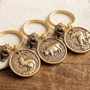 May include: A collection of gold-colored keychains. Each keychain features a round pendant with an animal design, including a tiger, bull, and cat. The keychains are arranged on a wooden surface, showcasing their intricate details and craftsmanship.