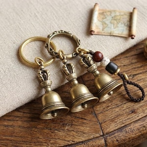 Könnte beinhalten: Drei antike Schlüsselanhänger mit Glocken aus Messing mit Kronendetails. Jede Glocke hat eine kleine Schlaufe zur Befestigung an einem Schlüsselring. Ein Schlüsselanhänger hat einen dekorativen Perlenakzent und eine braune Kordel. Eine aufgerollte Karte befindet sich im Hintergrund.