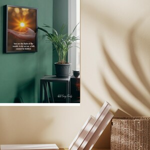 Puede incluir: Una impresión enmarcada con el texto "You are the light of the world. A city set on a hill cannot be hidden." La imagen dentro del marco representa una mano que sostiene un sol brillante. Una planta en maceta está sobre una mesa pequeña.