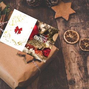 Puede incluir: Un regalo envuelto con una tarjeta de Navidad con un lazo rojo y detalles dorados. La tarjeta se coloca encima de un regalo envuelto con una ramita de pino y palitos de canela. Las palabras "Merry Christmas" son visibles.