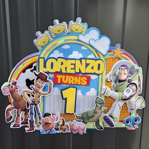 Puede incluir: Decoración de cumpleaños colorida con personajes de Toy Story. El diseño incluye a Woody, Buzz Lightyear y otros personajes, con el nombre "Lorenzo" y el número "1" destacados. Un arcoíris y nubes completan el aspecto festivo.