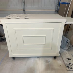 Op de afbeelding: Een witte, rollende bar met een rechthoekig ontwerp. De bar heeft een aanrechtblad met verschillende cirkelvormige uitsparingen en twee zijuitbreidingen. Het voorpaneel heeft een verzonken rechthoekig ontwerp. De bar staat op wielen.
