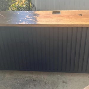 May include: A large wooden desk with a light brown top and a black, vertically-grooved front. The desk has a rectangular cutout and a circular hole on the top surface. The desk is on wheels.