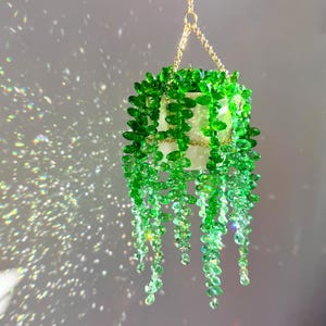 Peut inclure: Une suspension décorative représentant une plante en pot blanc avec des tiges tombantes composées de perles de cristal vert facettées. L'ensemble est suspendu par une fine chaîne en métal doré, créant des reflets lumineux sur les perles.