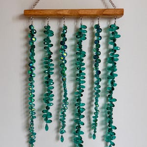 May include: A wall hanging featuring a horizontal wooden bar suspended by a silver chain. Six vertical strands of faceted, emerald green crystal beads hang from the bar, each ending in a single teardrop-shaped bead, creating a cascading effect.