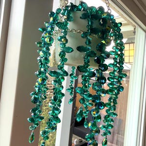 Peut inclure: Un ornement décoratif suspendu composé d'un petit pot blanc orné de nombreuses chaînes de perles en cristal vert émeraude à facettes. Les perles sont disposées pour imiter une plante tombante, suspendue par une structure en chaîne métallique argentée.