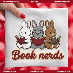 May include: White fabric featuring an embroidered design of three cartoon rabbits engrossed in books. The rabbits are white, grey, and brown. The text "Book nerds" is embroidered in burgundy beneath the rabbits.