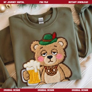 May include: Olive green sweatshirt featuring an embroidered bear wearing a green hat with a red feather, a pretzel necklace, and holding a mug of beer. The bear has pink cheeks and a sleepy expression. The text "MY JOURNEY DIGITAL" is at the top.