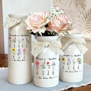 May include: Three white ceramic vases with floral designs and lace bows. The vases are decorated with the words "Grandma's Garden" and "Nana's Garden", along with floral illustrations and names. One vase holds pink roses.