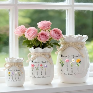 May include: Three white ceramic vases with frilled tops and rope detailing sit on a windowsill. The vases are adorned with floral illustrations and the text "Nana's Garden" and "Love Grows Here". One vase contains pink roses.