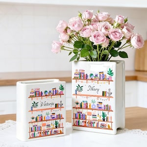 Op de afbeelding: Twee witte keramische vazen met een boekenplankontwerp. Eén vaas is gevuld met roze rozen. De vazen hebben de namen "Victoria" en "Mary" erop gedrukt. Het boekenplankontwerp bevat boeken, planten en globes.