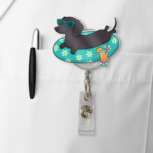 Puede incluir: Un perro salchicha negro con gafas de sol flota en un flotador de piscina turquesa con flores blancas y amarillas. El perro está en un carrete retráctil para insignias con un soporte de plástico transparente. Un bolígrafo negro está en el bolsillo de una bata blanca.