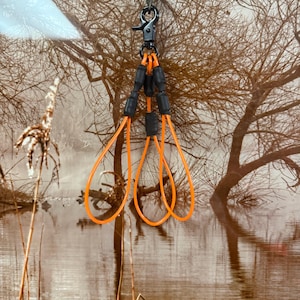 May include: A black metal clip with a green and brown braided cord is attached to a multi-strand lanyard. The lanyard features three black connectors and bright orange loops. The background shows a landscape with trees and water.