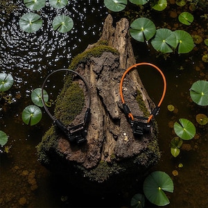 May include: Two bicycle cable locks, one brown and one orange, rest on a moss-covered tree stump in a pond with lily pads. The locks have black locking mechanisms and are designed for securing bicycles.