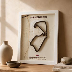 Könnte beinhalten: Ein gerahmtes Wandbild mit einem erhabenen, dunkel gefärbten 3D-Umriss der Silverstone-Rennstrecke. Der Druck enthält den Text BRITISH GRAND PRIX, SILVERSTONE CIRCUIT, OPENED: 1948, TURNS: 18, LENGTH: 5.891 KM, LAPS: 52 sowie eine kleine britische Flagge.