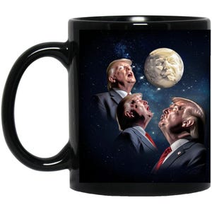May include: A black coffee mug with a white design of three men's faces looking up at a moon-like object. The men are wearing suits and ties. The background is a dark blue space with stars.