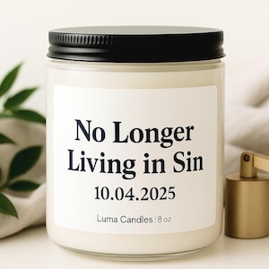 Peut inclure: Un pot de bougie en verre blanc avec un couvercle noir. L'étiquette indique "No Longer Living in Sin" et "10.04.2025". La bougie est de 227g et fabriquée par Luma Candles. Un objet doré se trouve à droite de la bougie.