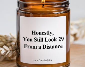 May include: A lit candle in a brown glass jar with a white label. The label reads "Honestly, You Still Look 29 From a Distance." The candle is from Luma Candles and is 8oz.