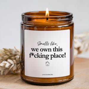 May include: An amber glass candle jar with a white label featuring the black text 'Smells like.. we own this f*cking place!' and a small house icon. The label also includes the text 'Luma Candles| 8oz'. A lit wick is visible at the top.