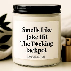 Peut inclure: Une bougie blanche dans un pot en verre transparent avec un couvercle noir. L'étiquette indique "Smells Like Jake Hit The F*cking Jackpot" avec "Luma Candles | 8oz" en dessous. Un flacon de parfum doré est visible en arrière-plan.