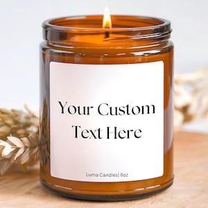 以下が含まれることがあります： 「Your Custom Text Here」と書かれた白いラベルが付いた、点灯した琥珀色のガラスのキャンドルジャー。キャンドルは8オンスで、背景には木製の表面があります。