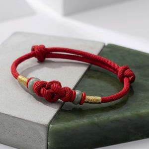 May include: Red rope bracelet with a central knot design and two light green beads. The bracelet features gold-colored accents and is displayed on a gray and green surface. A stylish accessory.