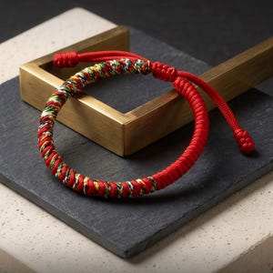 May include: A red and multicoloured woven bracelet. The bracelet has a vibrant design with red, yellow, green, and white threads. The adjustable closure is made of red cord. The bracelet is displayed on a dark grey surface.