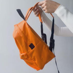 Może przedstawiać: Pomarańczowy plecak z czarnymi paskami i małą kieszenią. Plecak trzymany jest przez osobę ubraną w białą koszulę z długim rękawem. Torba ma prostokątny kształt i uchwyt.