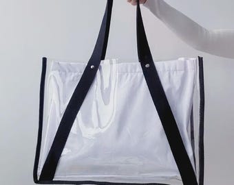 May include: Clear tote bag with black straps and trim. The bag has a white interior and is held by a person wearing a white long-sleeved shirt. The tote bag is designed for carrying items.