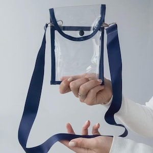 May include: A clear crossbody bag with a navy blue trim and strap. The bag has a rectangular shape with a flap closure and a navy blue strap that is approximately 1 inch wide. The bag is held up by a person.