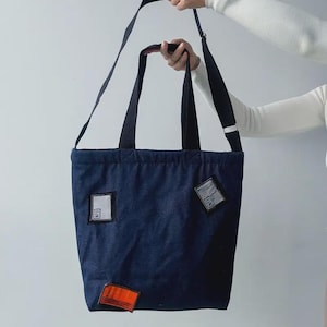 Puede incluir: Un bolso tote azul marino con correas y parches negros. El bolso tiene forma rectangular con una correa larga para el hombro y dos asas más cortas. Los parches son cuadrados y rectangulares, con detalles en blanco y naranja.