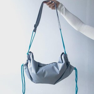 May include: A gray shoulder bag with a long strap and teal drawstrings. The bag is held up by a person's hand against a white background. The bag's shape is curved, with a zipper closure.
