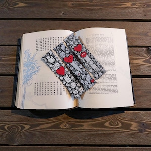 Peut inclure: Un livre ouvert avec trois marque-pages en tissu. Les marque-pages présentent un motif noir et blanc de visages souriants et de cœurs. Le livre est posé sur une surface en bois.