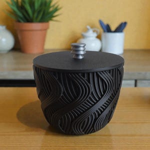 Peut inclure: Un récipient décoratif noir avec un couvercle et un motif texturé en forme de vagues. Le récipient est posé sur une surface en bois. Une petite plante dans un pot en terre cuite et d'autres objets décoratifs sont en arrière-plan.