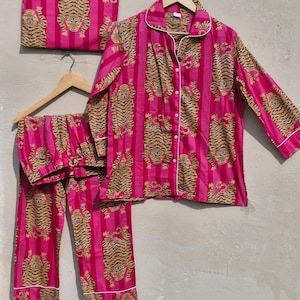 May include: A pink pajama set with a tiger print, including a button-down shirt, pants, and a matching storage bag. The set features vertical pink stripes and tiger illustrations in shades of brown and black. White piping trims the edges.