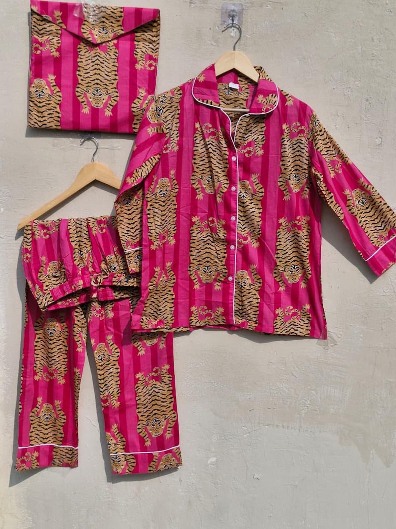 May include: A pink pajama set with a tiger print, including a button-down shirt, pants, and a matching storage bag. The set features vertical pink stripes and tiger illustrations in shades of brown and black. White piping trims the edges.