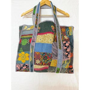 Op de afbeelding: Een patchwork tote bag met een verscheidenheid aan kleurrijke stoffen vierkanten. De tas heeft een bloemenpatroon, blauwe, gele en groene tinten en een zwart-witte gestreepte rand. De tas hangt aan een houten hanger.