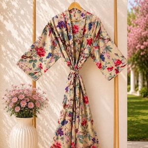 Puede incluir: Una bata de manga larga con un color base beige y un estampado floral vibrante en tonos rojos, azules y verdes. La prenda presenta un diseño cruzado con un cinturón de tela a juego en la cintura y mangas anchas y holgadas.