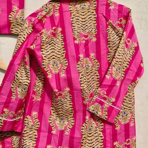 May include: Pink pajama set with a vertical stripe pattern and a repeating tiger design in shades of tan and black. The long-sleeved top and matching bottoms are trimmed with white piping.