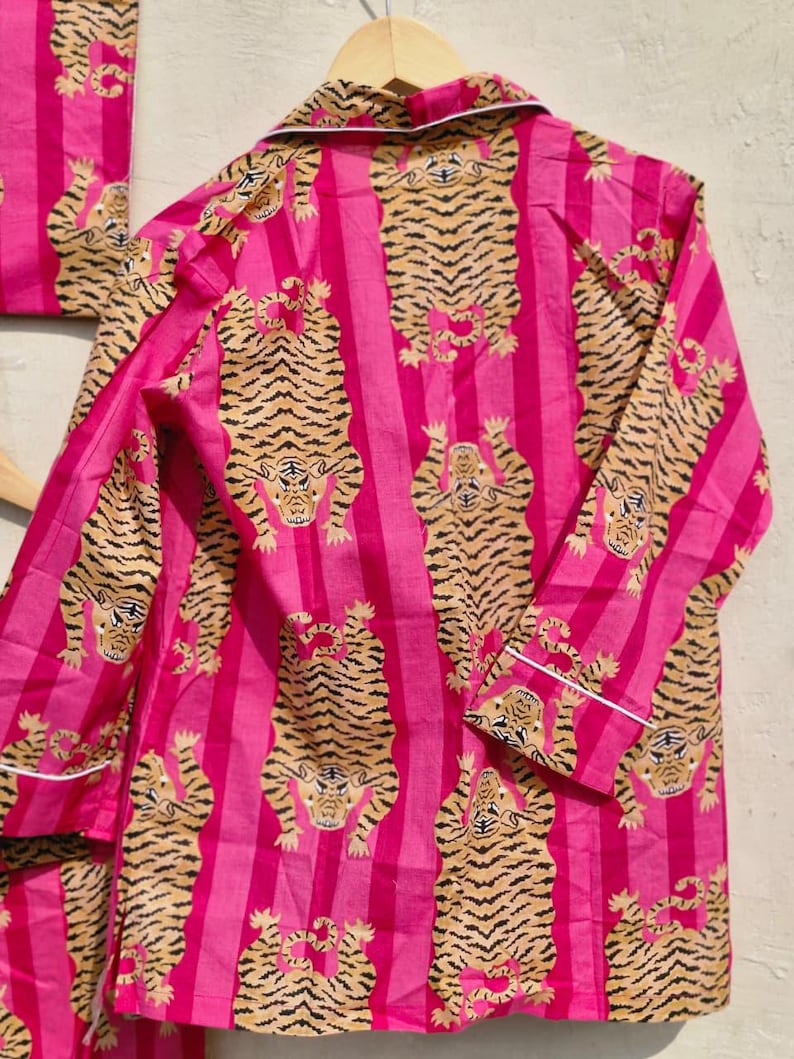 May include: Pink pajama set with a vertical stripe pattern and a repeating tiger design in shades of tan and black. The long-sleeved top and matching bottoms are trimmed with white piping.