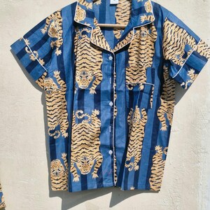 May include: Blue short-sleeved pajama top with a tiger print. The shirt has a button-down front, a notched collar, and white trim. The tiger pattern is in shades of tan and brown, set against blue vertical stripes.