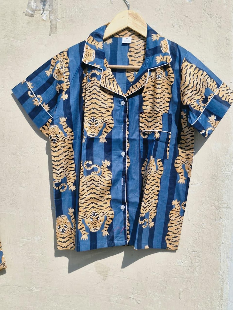 May include: Blue short-sleeved pajama top with a tiger print. The shirt has a button-down front, a notched collar, and white trim. The tiger pattern is in shades of tan and brown, set against blue vertical stripes.