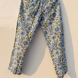 May include: Light blue and white floral print pajama pants hanging on a wooden hanger. The pants feature a repeating pattern of blue flowers and green leaves on a white background. The pants have an elastic waistband.
