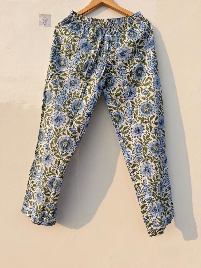 May include: Light blue and white floral print pajama pants hanging on a wooden hanger. The pants feature a repeating pattern of blue flowers and green leaves on a white background. The pants have an elastic waistband.