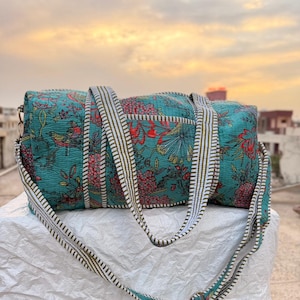 Könnte beinhalten: Eine türkisblaue Reisetasche mit einem Blumenmuster in Rot, Gelb und Grün. Die Tasche hat einen gestreiften Riemen in Weiß und Olivgrün. Die Tasche hat einen Durchmesser von ca. 25 cm und ist 50 cm lang.