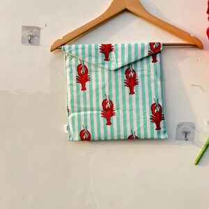 May include: A square fabric item with a red lobster pattern on a light green and white striped background. The item is folded and hanging on a wooden hanger. The lobster design is repeated across the fabric.