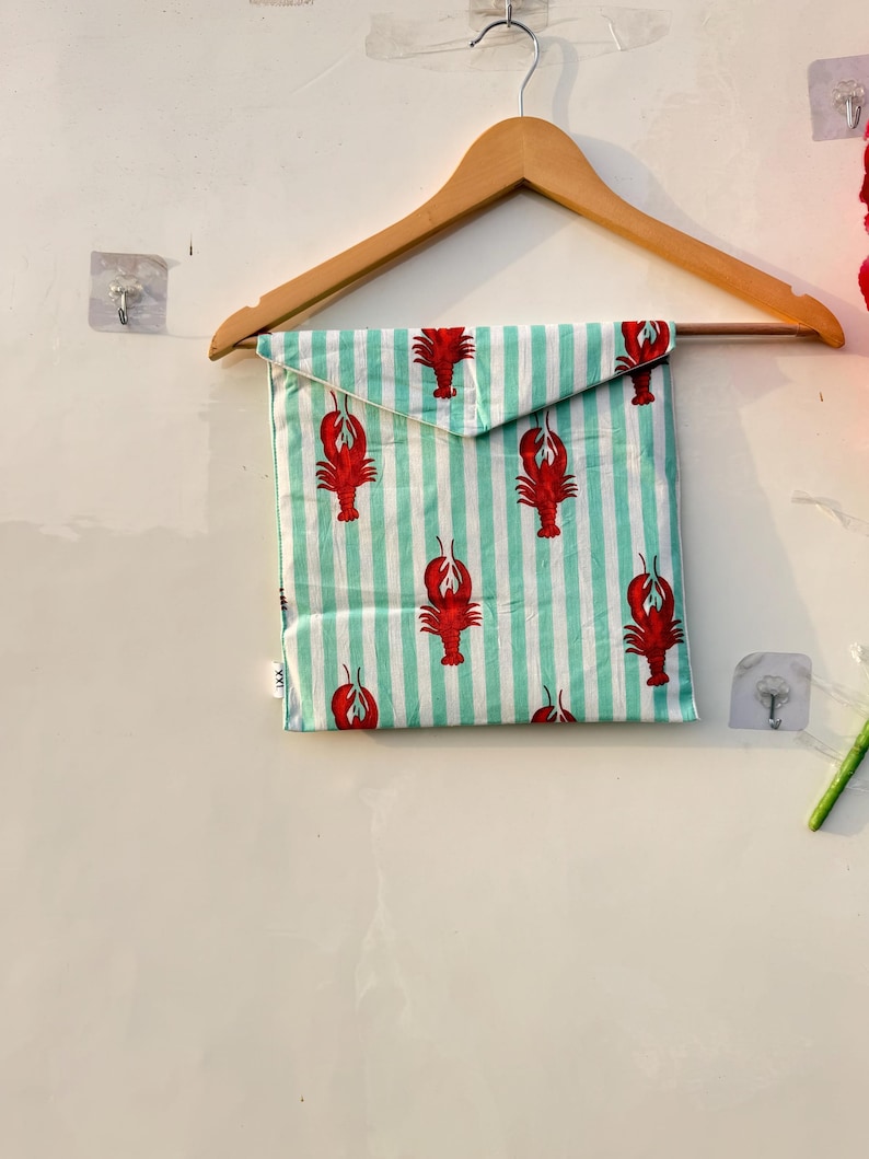 May include: A square fabric item with a red lobster pattern on a light green and white striped background. The item is folded and hanging on a wooden hanger. The lobster design is repeated across the fabric.