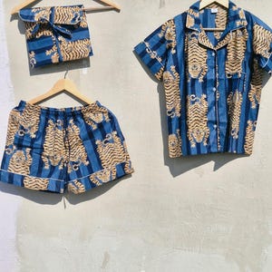 May include: A blue pajama set with a tiger print, comprising a short-sleeved shirt, shorts, and a pouch. The fabric displays vertical blue stripes and a repeating pattern of tan tigers. The shirt and shorts are trimmed with white. The set is hung on wooden hangers.