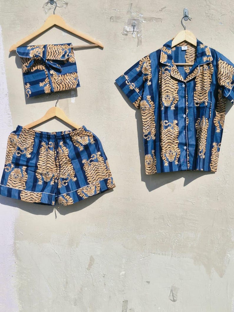 May include: A blue pajama set with a tiger print, comprising a short-sleeved shirt, shorts, and a pouch. The fabric displays vertical blue stripes and a repeating pattern of tan tigers. The shirt and shorts are trimmed with white. The set is hung on wooden hangers.