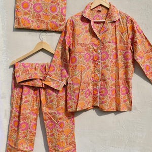 May include: A peach-colored pajama set with a floral pattern in shades of orange and pink. The set includes a button-down shirt, pants, and a matching pouch. The shirt and pants have white trim. The pajamas are hanging on wooden hangers.